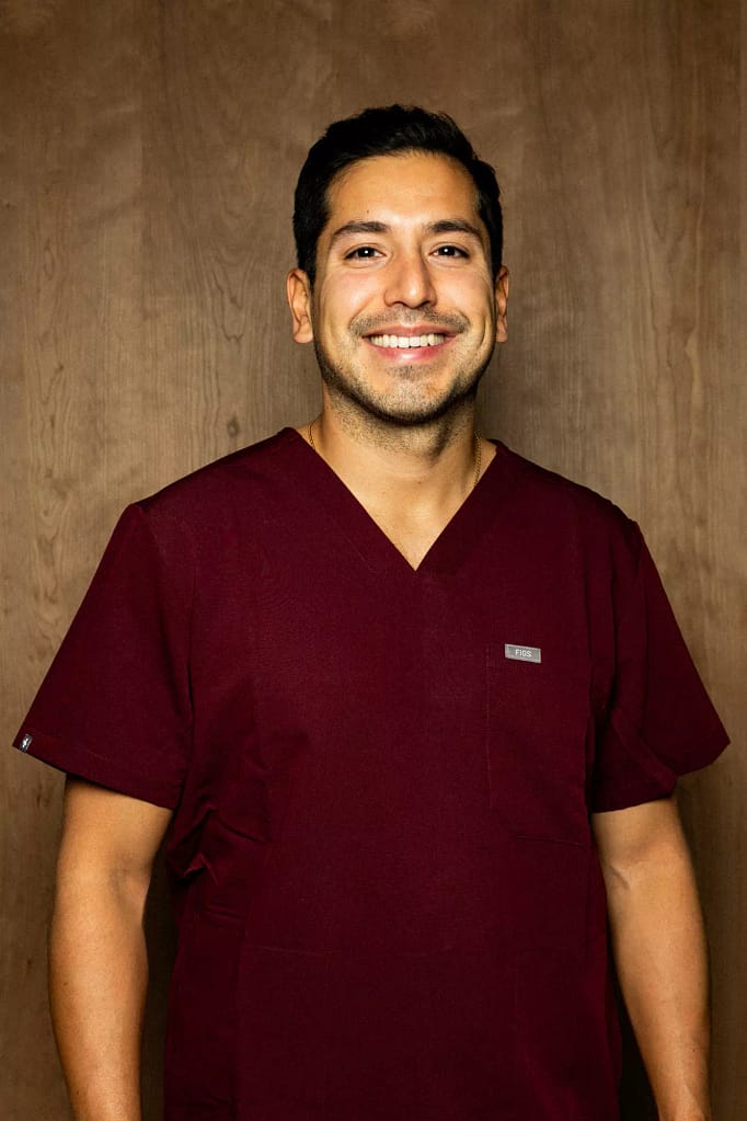 Portrait du Dr Mauricio Pinto, en tenue médicale, souriant, devant un fond en bois.