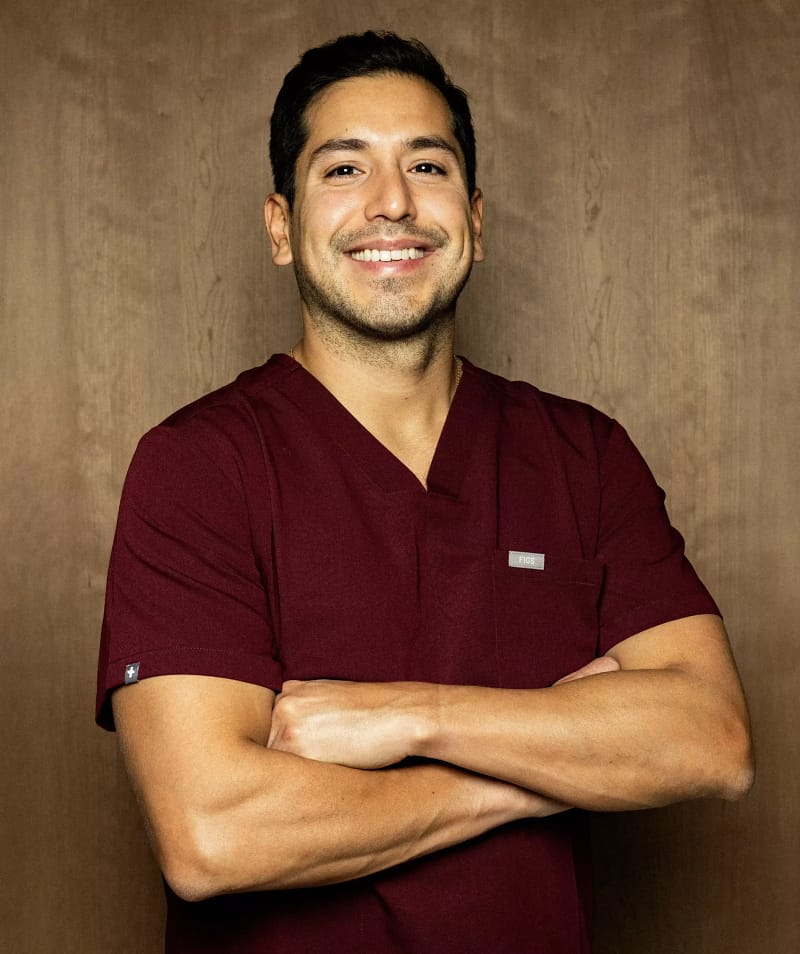 Portrait du Dr Mauricio Pinto, en tenue médicale, les bras croisés, souriant, devant un fond en bois.